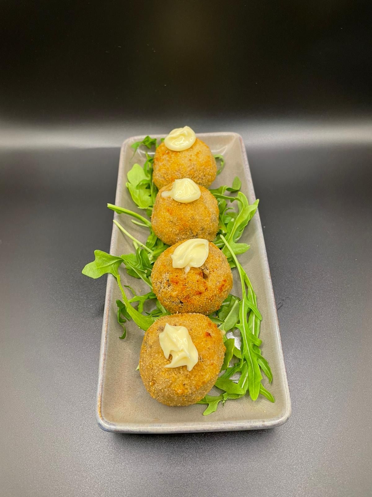 Four golden arancini balls with aioli and rocket on a slate plate, dark background