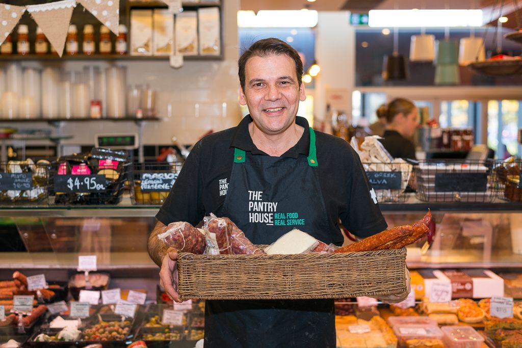 Smiling Pantry House owner in branded apron holding deli goods inside the store with full deli counter visible