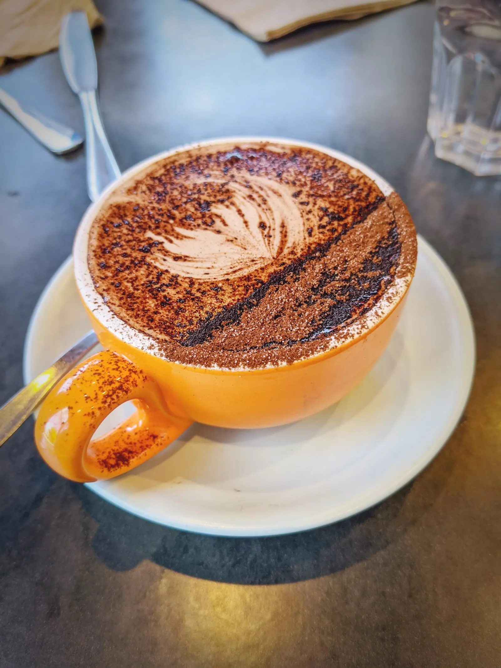 Mocha in an orange mug with cocoa-dusted latte art on a dark metal café table