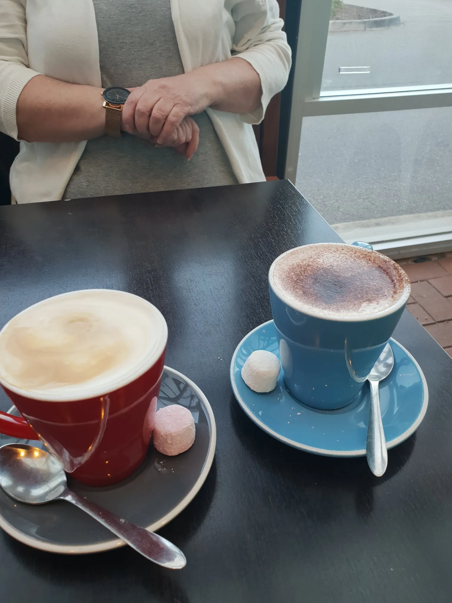 Two colourful coffee mugs on a café table with a patron's hands and rainy window view in background