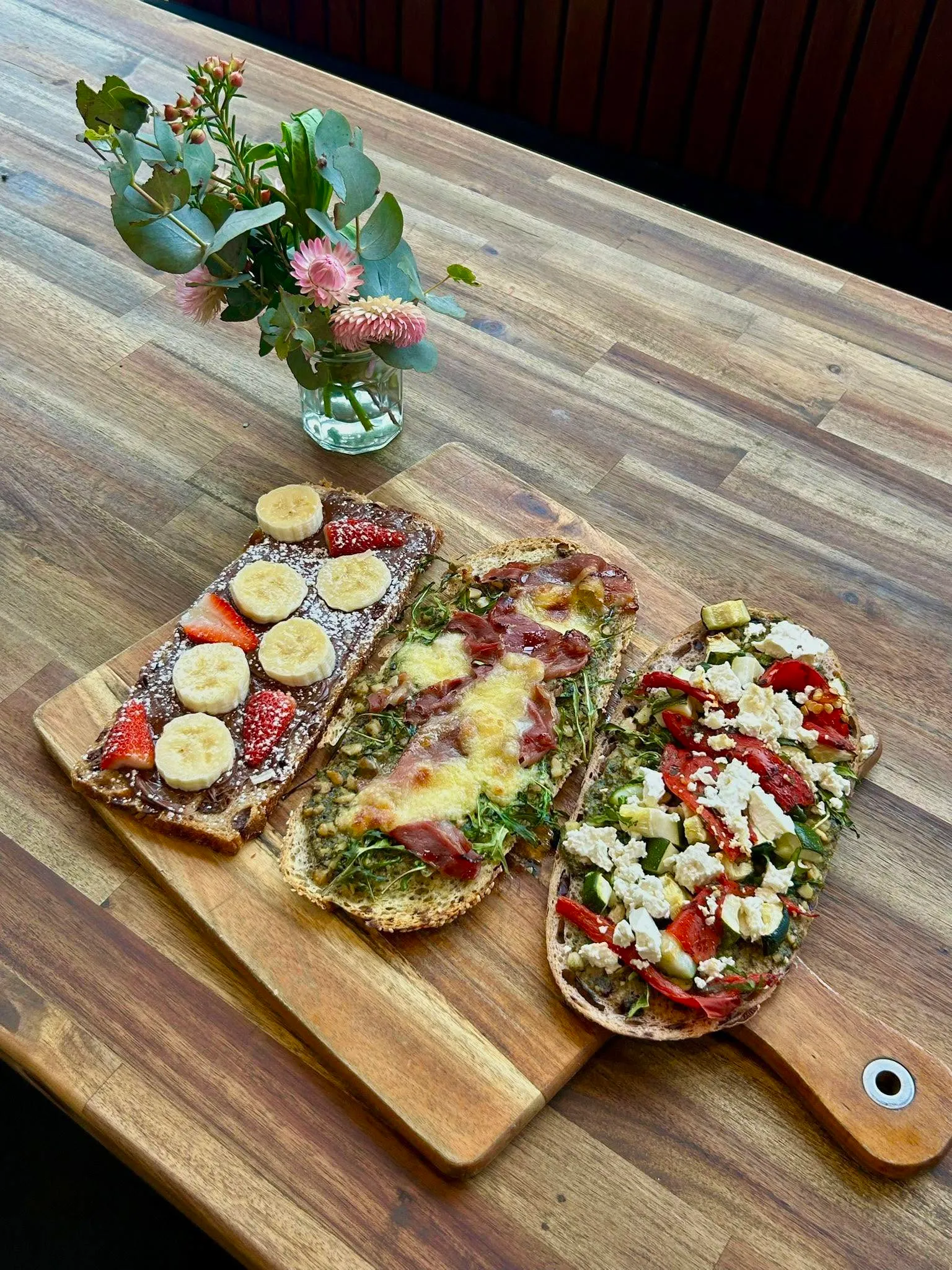 Three open-faced toasts on a wooden board — sweet banana, cheese herb, and white bean feta varieties
